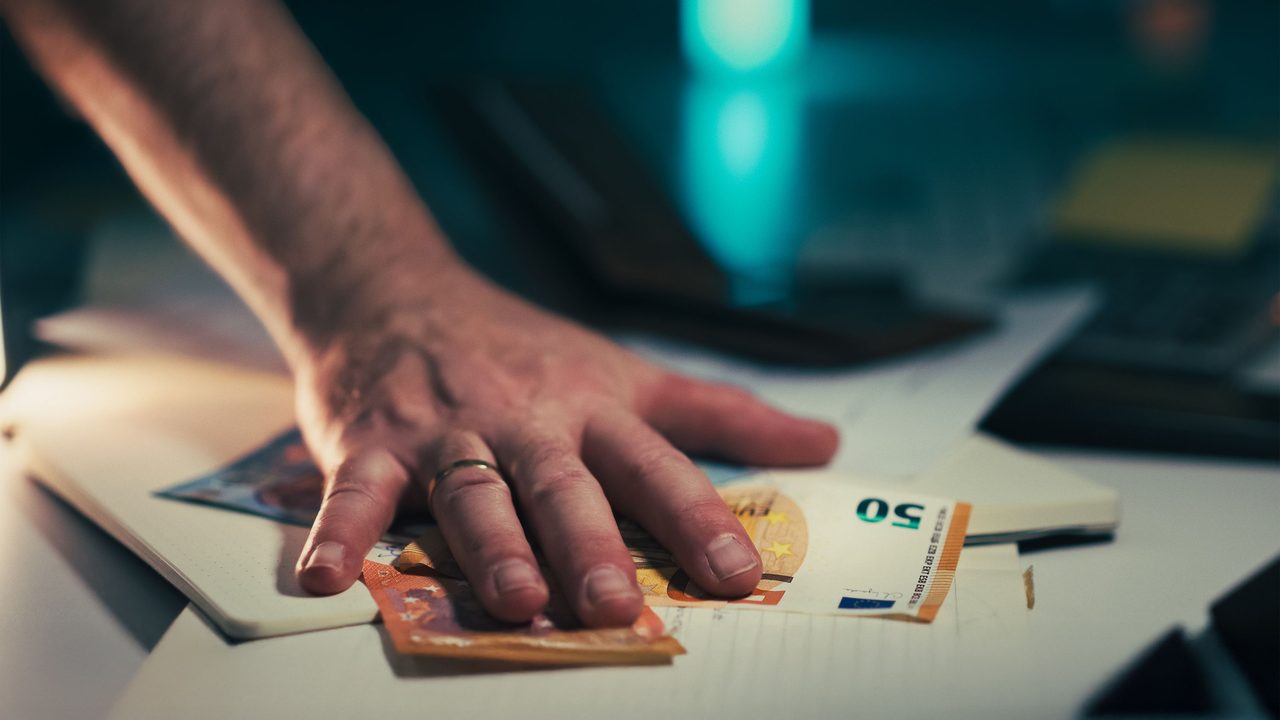 Een hand grijpt eurobiljetten van een bureau in schemerig licht, symbool voor het fortuin van een oud-topvoetballer